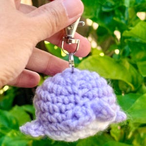 May include: A small, round, lavender and white crocheted whale keychain. The plush, textured yarn figure features a rounded body with small side fins and is attached to a silver-toned metal swivel lobster clasp by a sturdy metal jump ring.