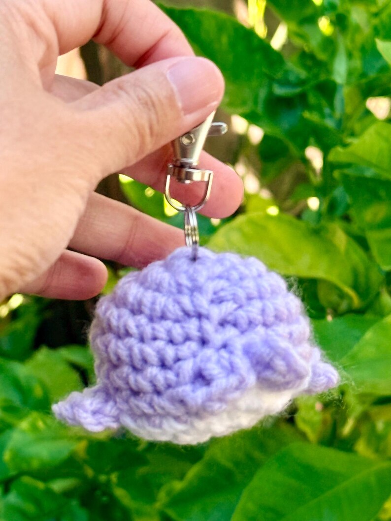 May include: A small, round, lavender and white crocheted whale keychain. The plush, textured yarn figure features a rounded body with small side fins and is attached to a silver-toned metal swivel lobster clasp by a sturdy metal jump ring.