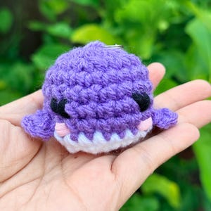 May include: A small, round, purple crochet whale keychain featuring black embroidered eyes, light pink stitched cheeks, and a white yarn mouth. The plush toy has two small side fins and a metal ring attached to the top for hanging purposes.