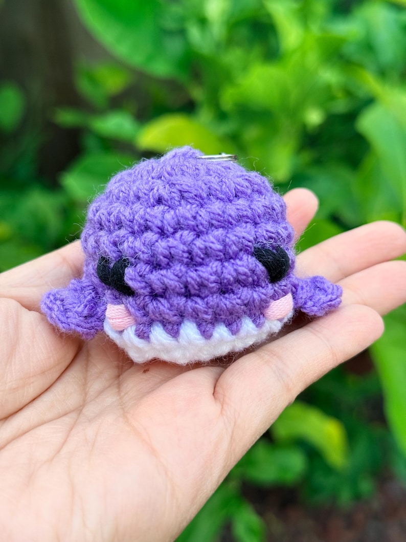 May include: A small, round, purple crochet whale keychain featuring black embroidered eyes, light pink stitched cheeks, and a white yarn mouth. The plush toy has two small side fins and a metal ring attached to the top for hanging purposes.