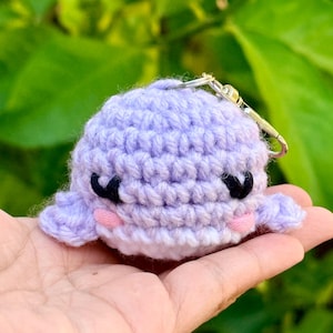 May include: A small, round, light purple crocheted whale keychain. The whale features black yarn embroidered eyes, small pink yarn circles for cheeks, and a silver-toned metal keyring attached to the top of its head with a lobster clasp.