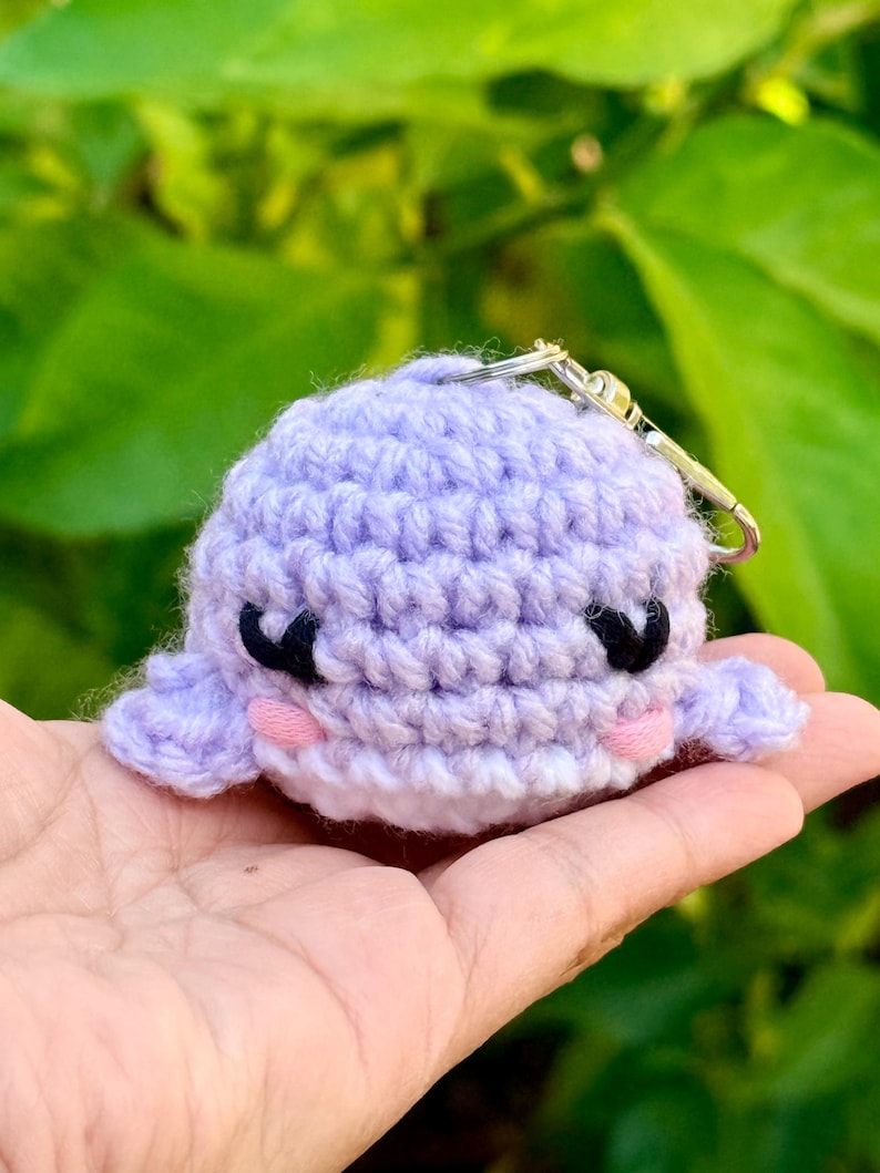 May include: A small, round, light purple crocheted whale keychain. The whale features black yarn embroidered eyes, small pink yarn circles for cheeks, and a silver-toned metal keyring attached to the top of its head with a lobster clasp.