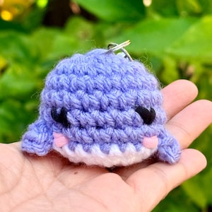 May include: A small, light purple crocheted whale keychain featuring a white underbelly, black embroidered eyes, and small pink stitched cheeks. The plush toy is crafted with visible yarn loops and includes a silver metal keyring at the top.