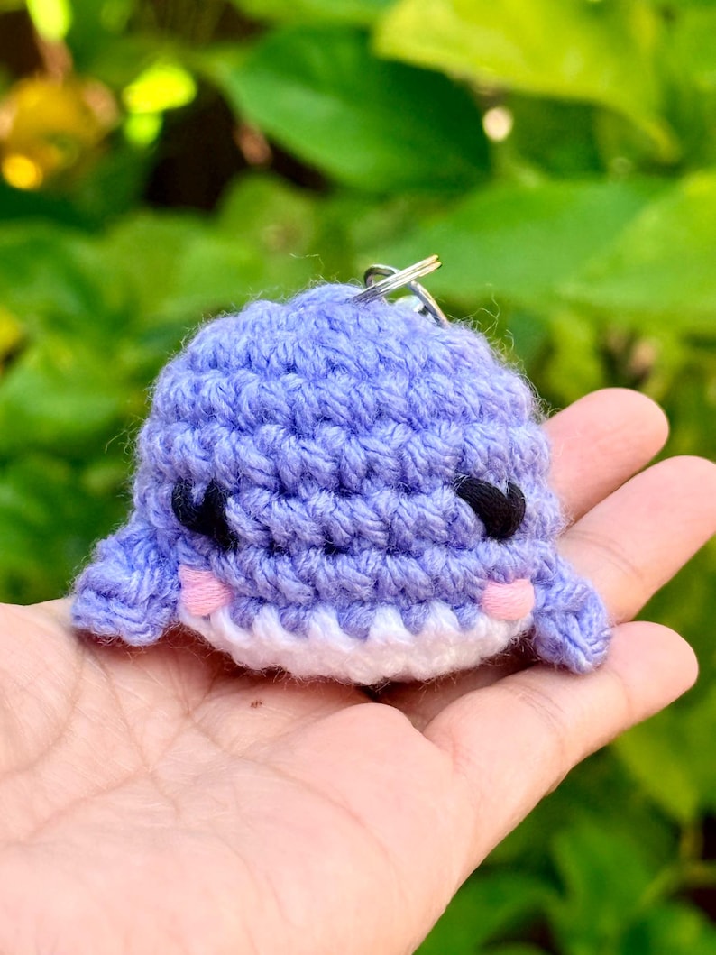 May include: A small, light purple crocheted whale keychain featuring a white underbelly, black embroidered eyes, and small pink stitched cheeks. The plush toy is crafted with visible yarn loops and includes a silver metal keyring at the top.