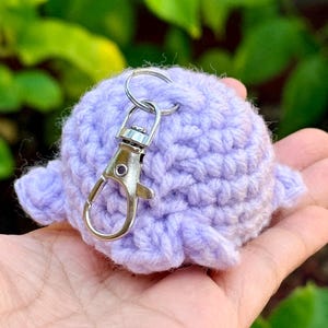 May include: A light purple, round, crocheted yarn keychain featuring a small, textured, stuffed sphere with attached side fins and a silver-toned metal lobster claw clasp attached to a circular ring at the top of the fiber-based object.
