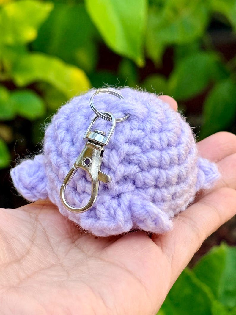 May include: A light purple, round, crocheted yarn keychain featuring a small, textured, stuffed sphere with attached side fins and a silver-toned metal lobster claw clasp attached to a circular ring at the top of the fiber-based object.