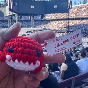 May include: A red crocheted plush toy with black eyes and a white mouth, attached to a rectangular white tag that reads HI ARMY! I'M KORE-RANG! MAY 16-17, 19, 2026 STANFORD STADIUM with the BTS WORLD TOUR logo and three horizontal lines.