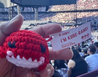 May include: A red crocheted plush toy with black eyes and a white mouth, attached to a rectangular white tag that reads HI ARMY! I'M KORE-RANG! MAY 16-17, 19, 2026 STANFORD STADIUM with the BTS WORLD TOUR logo and three horizontal lines.