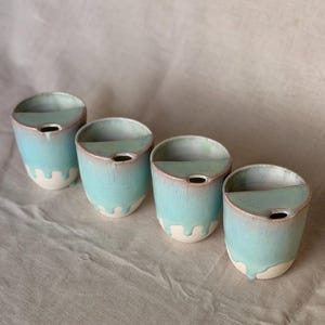May include: Four ceramic cups with a semi-circular interior shelf and a small circular opening. The exterior features a light blue glaze that drips down towards the base, revealing a cream-coloured ceramic body with pinkish-brown rim accents.