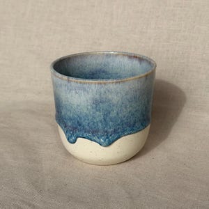 May include: A ceramic mug featuring a two-tone design with a speckled cream-coloured base and a glossy, deep blue glaze that drips down from the rim. The interior of the mug is finished in a smooth, matching blue glaze with a wide, open top.
