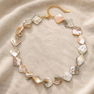 May include: A necklace featuring a series of iridescent, square-shaped shell beads in shades of cream, peach, and grey. The beads are linked together with gold-toned metal hardware, including a lobster claw clasp and an adjustable chain extender.