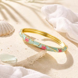 May include: A gold-tone hinged bangle bracelet featuring a light blue enamel finish with a repeating pattern of small pink starfish and swirling wave-like lines. The bracelet has a polished metallic interior and a secure clasp mechanism.