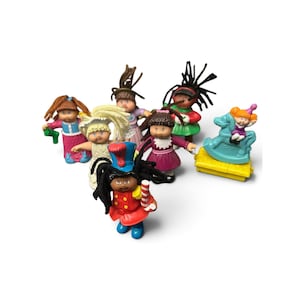 May include: A collection of seven small plastic Cabbage Patch Kids figurines featuring various outfits, including a soldier with a candy cane, a child on a rocking horse, and others in colorful dresses, all with yarn hair and distinct facial expressions.