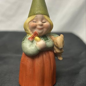 May include: A ceramic figurine of a person wearing a tall olive green pointed hat, a light green long-sleeved top with dark green accents, and a solid orange skirt. The figure has blonde hair, closed eyes, and holds a small tan animal.
