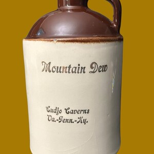 May include: A ceramic jug with a brown glazed top and a cream-colored body. The text Mountain Dew is printed in brown, followed by the text Cudjo Caverns Va.-Tenn.-Ky. The jug features a single handle on the side and a narrow neck opening.