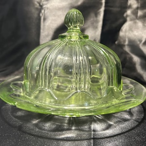 May include: A translucent light green glass butter dish featuring a domed lid with vertical ribbing and a small knob handle. The base is a circular plate with a scalloped edge design, providing a clear view of the pressed glass construction.