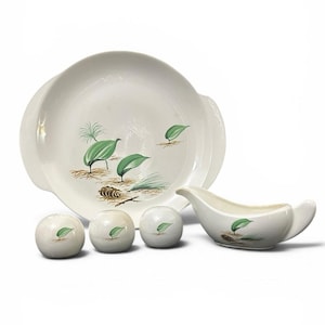 May include: A white ceramic serving set featuring a large round platter, a gravy boat, and three spherical shakers. Each piece is decorated with a hand-painted motif of green leaves, brown pinecones, and thin brown twigs on a white background.