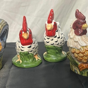 May include: A set of four ceramic rooster figurines featuring detailed plumage in shades of red, yellow, white, and blue. Each rooster is depicted standing on a green base, with two figures facing forward and two figures facing to the side.