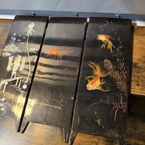 May include: Three rectangular black lacquered panels featuring hand-painted designs. The left panel shows a figure in a conical hat near a palm tree. The center and right panels depict orange goldfish swimming among aquatic plants and seaweed.