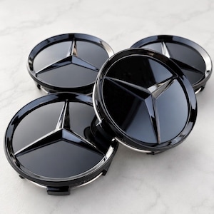 May include: A set of four circular wheel center caps featuring a glossy black finish and a prominent silver three-pointed star emblem in the center. Each cap has a raised, polished metallic star design set against a smooth, dark, reflective surface.