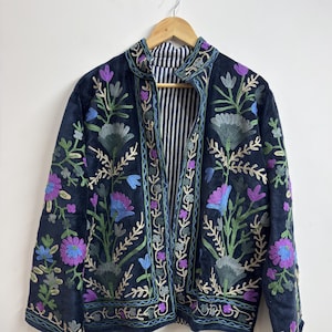Peut inclure: Une veste bleu marine foncé ornée de broderies florales complexes dans des tons de violet, bleu clair et vert. La veste présente un col montant, des manches longues et une ouverture frontale révélant une doublure à rayures verticales noires et blanches.