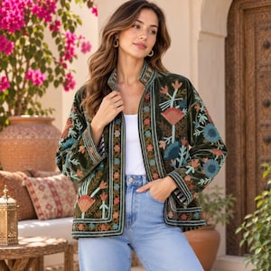 Könnte beinhalten: Eine olivgrüne Samtjacke mit langen Ärmeln und offener Vorderseite. Das Kleidungsstück ist mit aufwendigen Blumenstickereien in Orange-, Blau- und Petroltönen entlang der Vorderteile, Ärmelbündchen und des Saums verziert und wird über einem weißen Oberteil getragen.