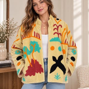 Könnte beinhalten: Eine hellgelbe Cordjacke mit Stehkragen und langen Ärmeln. Die Jacke ist mit bunten Stickereien verziert, darunter türkisfarbene Bäume, rotes Blattwerk, orangefarbene Kreise und eine große, schwarze, vogelähnliche Silhouette.