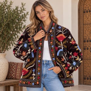 Puede incluir: Una chaqueta de terciopelo marrón oscuro con bordados florales multicolores vibrantes en la parte delantera y las mangas. La chaqueta tiene cuello recto, mangas largas y un diseño abierto, usada sobre una blusa blanca y jeans azules.