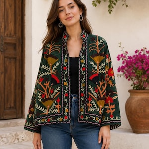 May include: A dark green open-front jacket featuring intricate multi-colored floral embroidery in shades of red, orange, and yellow. The garment has long sleeves, a mandarin-style collar, and a straight hemline, worn over a black top and blue jeans.