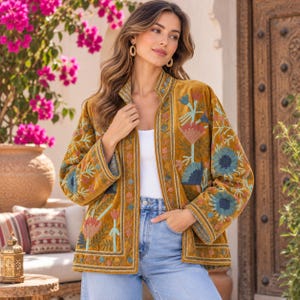 Könnte beinhalten: Eine senfgelbe Samtjacke mit offener Vorderseite und Stehkragen. Das Kleidungsstück ist mit aufwendigen Blumenstickereien in Blau-, Rosa- und Grüntönen verziert, die die Ärmel und Vorderteile über einem weißen Oberteil bedecken.