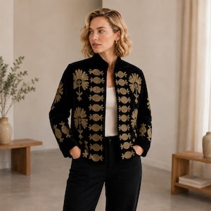 Puede incluir: Una chaqueta negra de manga larga con cuello alto y bordados en tono dorado. Los paneles frontales y las mangas están decorados con motivos florales y circulares repetitivos. La chaqueta se lleva sobre una blusa blanca y pantalones negros.