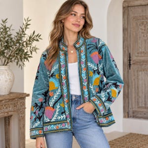 Könnte beinhalten: Eine petrolfarbene Samtjacke mit Stehkragen und langen Ärmeln. Die Jacke ist mit lebendigen, mehrfarbigen Blumenstickereien in Orange-, Gelb-, Rosa- und Hellblautönen verziert, die in vertikalen Bahnen und Mustern angeordnet sind.