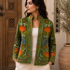 Puede incluir: Una chaqueta de terciopelo verde oliva con cuello mao y frente abierto. La chaqueta está decorada con intrincados bordados en tonos naranja, azul y verde claro, que representan patrones florales y botánicos en los paneles frontales.