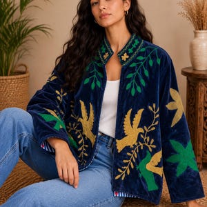 Puede incluir: Una chaqueta de terciopelo azul marino con bordados coloridos de pájaros dorados y hojas verdes. La chaqueta tiene cuello mao, mangas largas y un diseño abierto en la parte delantera. Se lleva sobre una camiseta blanca y vaqueros azules.