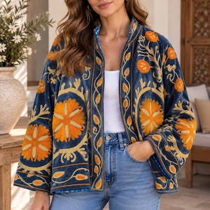 Op de afbeelding: Een marineblauw fluwelen jasje met levendige oranje en gele bloemenborduursels. Het jasje heeft een opstaande kraag, wijde mouwen en een open voorzijde. Het wordt gedragen over een effen wit topje en een lichtblauwe spijkerbroek.
