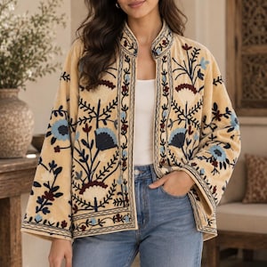 Può includere: Una giacca beige aperta sul davanti con colletto alla coreana e maniche lunghe e ampie. Il capo è decorato con intricati ricami floreali nei toni del rosso scuro, azzurro e blu navy, con una canotta bianca e jeans blu.