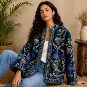 Può includere: Una giacca in velluto blu scuro con apertura frontale e maniche lunghe, decorata con vivaci ricami floreali nei toni del viola, azzurro e verde. Il design presenta un colletto alla coreana e intricati motivi botanici su tutto il capo.