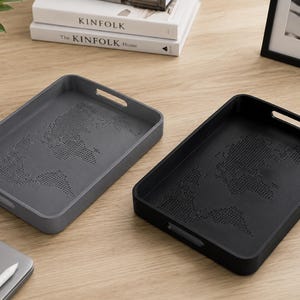 May include: Two rectangular serving trays, one in dark gray and one in black, each featuring a dotted world map design embossed on the base. Both trays have integrated handles on the short sides and raised edges for containment.