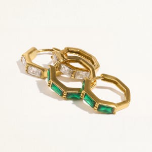 May include: A set of three gold-toned hoop earrings with a hexagonal shape. Two earrings feature clear rectangular baguette-cut stones, while the third earring is set with vibrant green rectangular baguette-cut stones along the outer edge.