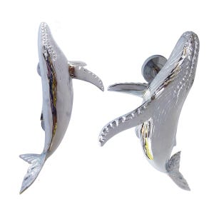 May include: Two silver-toned metal cabinet knobs shaped like humpback whales. Each knob features a polished, reflective surface with detailed textures on the fins and body, and a circular mounting base attached to the back of each whale figure.