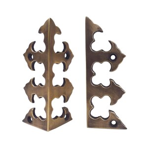 May include: Two antique brass corner protectors featuring an ornate, cut-out design with multiple points and pre-drilled screw holes. These metal hardware pieces have a dark, aged finish and are shaped to fit securely around the edges of furniture.