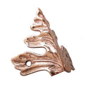 May include: A metallic copper-colored corner bracket featuring an intricate, embossed leaf-like design with a textured surface. The hardware includes two visible circular screw holes for mounting, with a curved, organic shape and a polished finish.