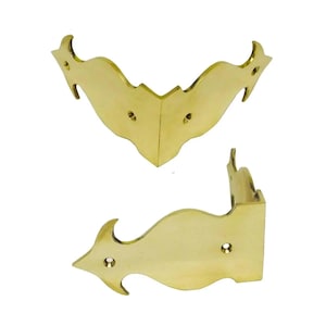 May include: Two polished brass corner protectors featuring a curved, decorative silhouette with pointed ends and pre-drilled screw holes. These metal hardware pieces are designed to fit over the edges of furniture or boxes to provide reinforcement.