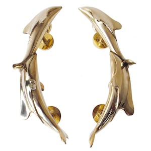 May include: A pair of polished gold-toned metal door handles shaped like leaping dolphins. Each handle features a curved, elongated body with smooth, reflective surfaces and two circular mounting bases for attachment to a door surface.