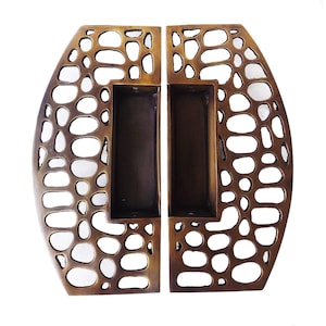 May include: A pair of bronze-toned metal door handles featuring a cutout design with irregular, pebble-like shapes. Each handle has a rectangular recessed grip area in the center, with visible screw holes at the top and bottom of the inner cavity.