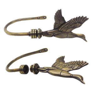 Puede incluir: Un par de alzapaños para cortinas de metal en tono latón antiguo con un diseño esculpido de un pato en vuelo. Un alzapaño se muestra ensamblado, mientras que el otro está separado en la conexión roscada, revelando la pieza interna.
