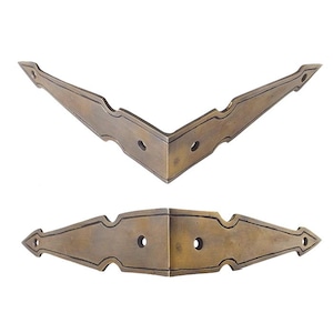 Op de afbeelding: Twee V-vormige hoekbeschermers van messing met een donkere, verouderde patina-afwerking. Elk stuk heeft een puntig, hoekig ontwerp met decoratieve uitsparingen langs de randen en twee voorgeboorde ronde gaten voor montage op meubels of kisten.