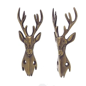 May include: Two identical brass-toned metal wall hooks shaped like deer heads with antlers. Each hook features a sculpted stag head with prominent ears and branching antlers, including two circular mounting holes at the base for attachment to a wall.