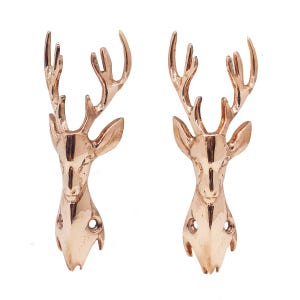 May include: A pair of matching wall hooks shaped like deer heads with antlers, finished in a polished rose gold metallic tone. Each hook features two circular screw holes at the base for mounting, with a smooth, reflective surface texture.