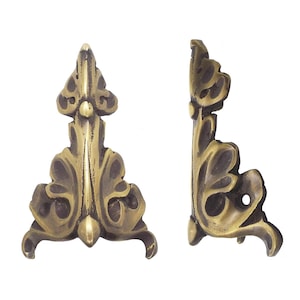 May include: Two brass corner protectors featuring an ornate, symmetrical leaf-like design with a dark, antiqued finish. The metal pieces are shaped to fit onto the edges of an object, with one shown from the front and one from the side profile.